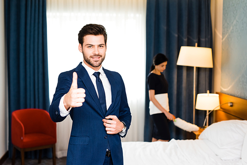 selective focus of happy receptionist showing thumb up near maid in hotel room