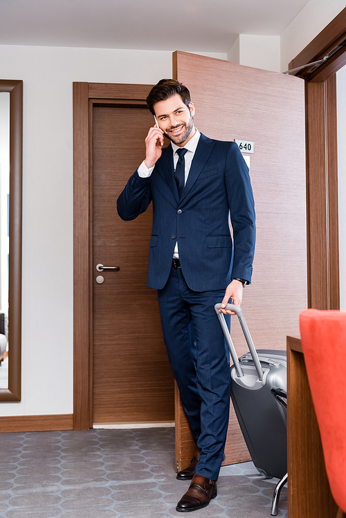 cheerful man in suit talking on smartphone and holding luggage