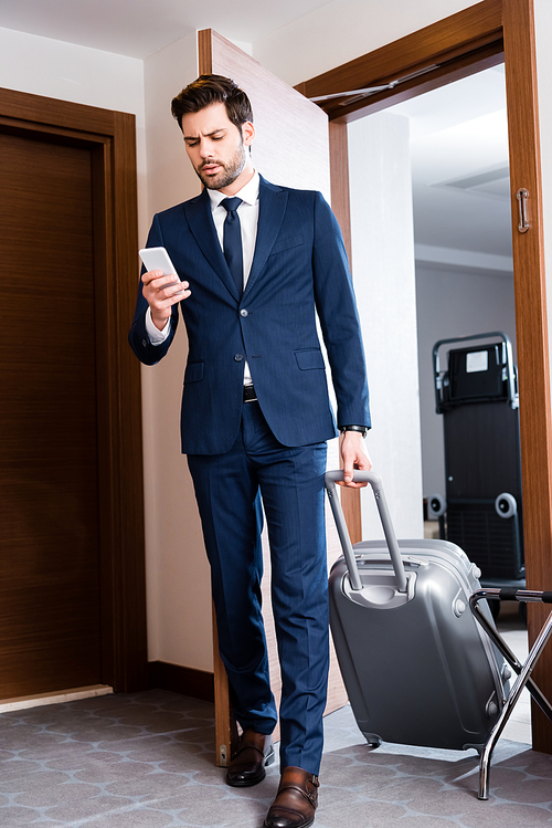 handsome businessman looking at smartphone while standing with suitcase in hotel