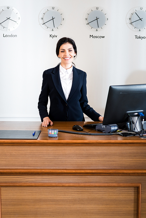 cheerful brunette receptionist in formal wear standing near computer monitor in hotel
