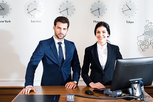 cheerful woman near serious receptionist in suit standing at reception desk