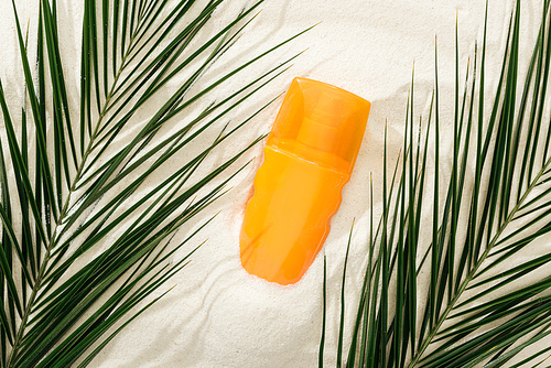 orange bottle of sunscreen on sand with green palm leaves