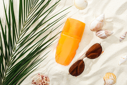 orange bottle of sunscreen on sand with seashells, green palm leaf and stylish sunglasses