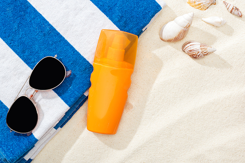 orange bottle of sunscreen on sand with seashells, striped towel and sunglasses