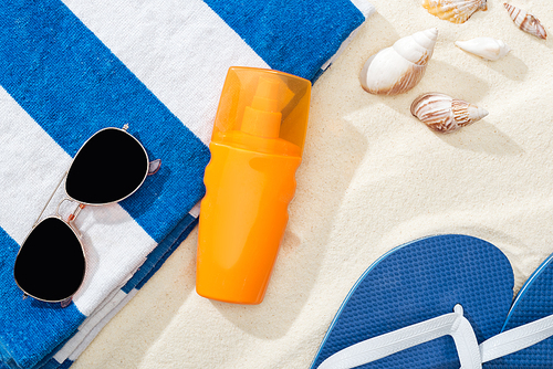 orange bottle of sunscreen on sand near striped towel, blue flip flops, sunglasses and seashells