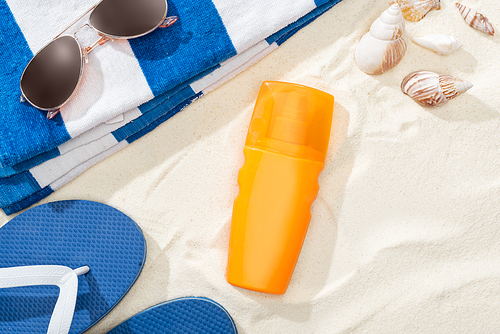 bottle of sunscreen on sand near striped towel, blue flip flops, sunglasses and seashells