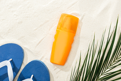 orange sunscreen near green palm leaf on sand with blue flip flops