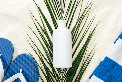 white sunscreen lotion on green palm leaf on sand with blue flip flops and towel