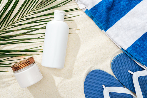 white sunscreen lotion and cream near green palm leaf on sand with blue flip flops and towel