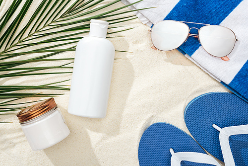 white sunscreen lotion and cream near green palm leaf on sand with blue flip flops, sunglasses and towel