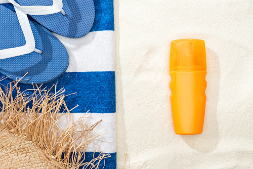 top view of sunscreen, straw hat, flip flops and striped blue and white towel on golden sand