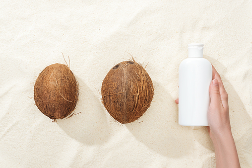 cropped view of woman holding white sunscreen lotion near coconuts on golden sand