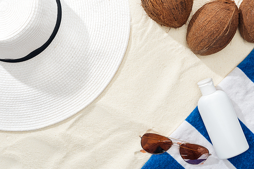 top view of sunglasses, striped towel, coconuts and white straw hat near white lotion on sand