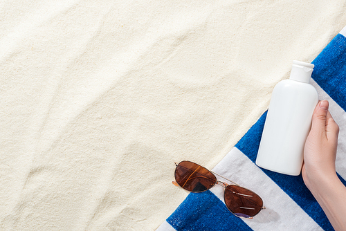 cropped view of woman holding sunscreen in hand near striped towel and sunglasses on sand