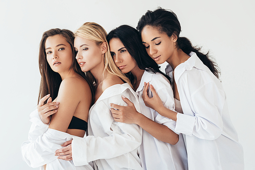 four attractive multiethnic women in white shirts embracing isolated on grey