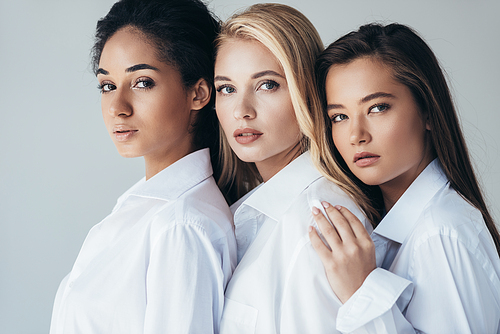 three attractive multiethnic girls in white shirts embracing isolated on grey