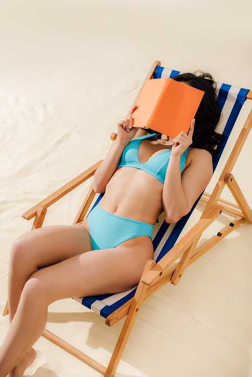 girl in bikini lying on deck chair and covering face with book on beach