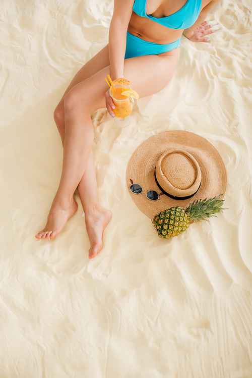 top view of woman in bikini with cocktail, straw hat, pineapple and sunglasses on beach