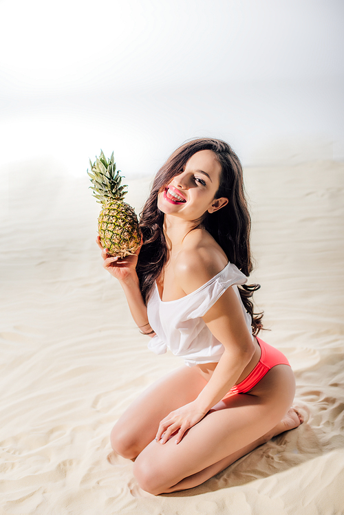 beautiful smiling girl sitting, Looking At Camera and posing with pineapple on sandy beach