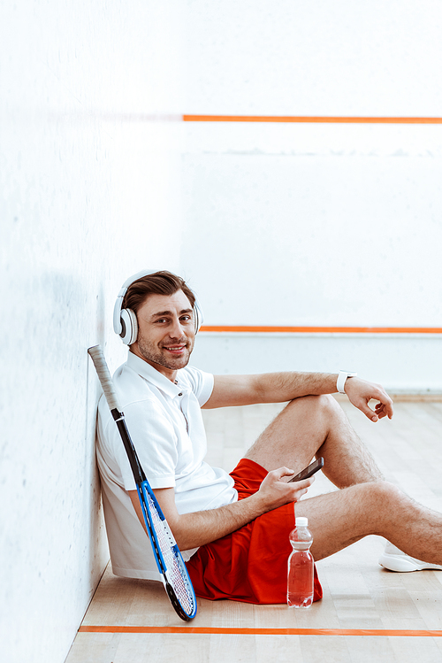 Smiling squash player listening music in headphones and using smartphone