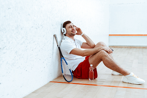 Smiling squash player sitting on floor and listening music in headphones