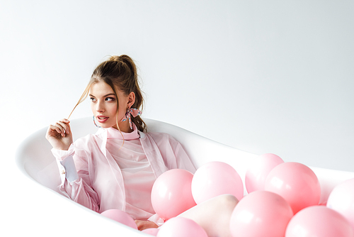 attractive girl touching hair while lying in bathtub with pink air balloons on white