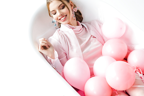 cheerful young woman lying in bathtub with pink air balloons on white