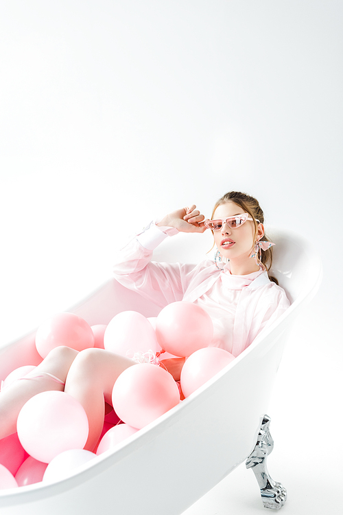 stylish young woman lying in bathtub with pink air balloons and touching sunglasses on white
