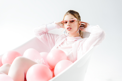 stylish young woman in sunglasses lying in bathtub with pink air balloons on white