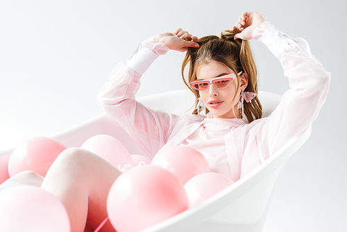 stylish young woman in sunglasses lying in bathtub with pink air balloons and touching hair on white