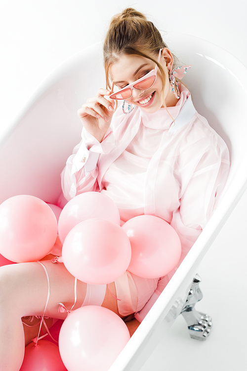overhead view of cheerful girl touching sunglasses while lying in bathtub with pink air balloons on white