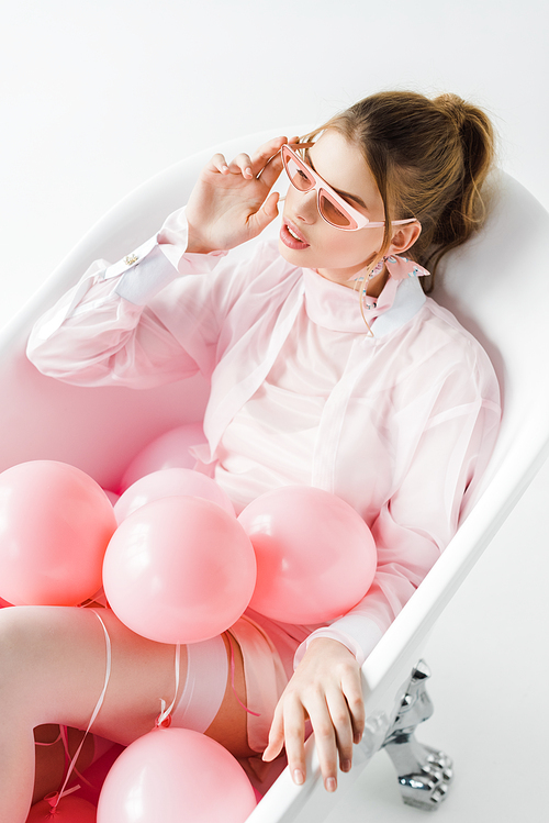overhead view of attractive girl touching sunglasses while lying in bathtub with pink air balloons on white