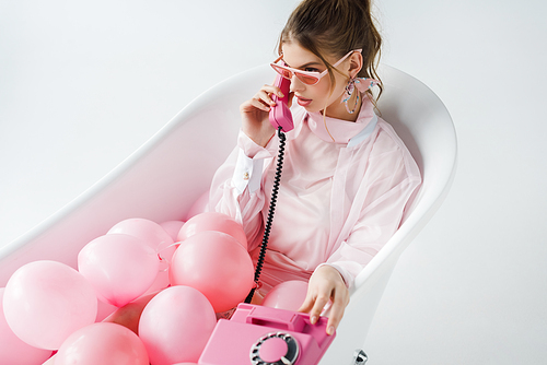 attractive girl in sunglasses talking on retro phone while lying in bathtub with pink air balloons on white