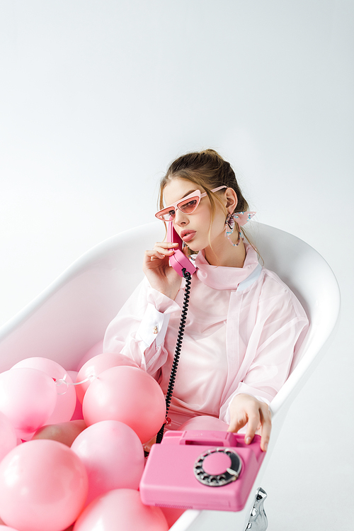 girl in sunglasses talking on retro phone while lying in bathtub with pink air balloons on white