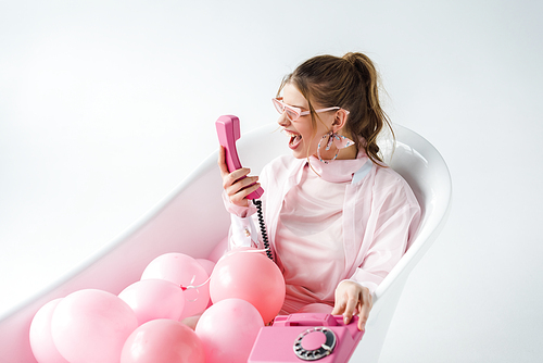 girl in sunglasses screaming at pink retro phone while lying in bathtub with air balloons on white