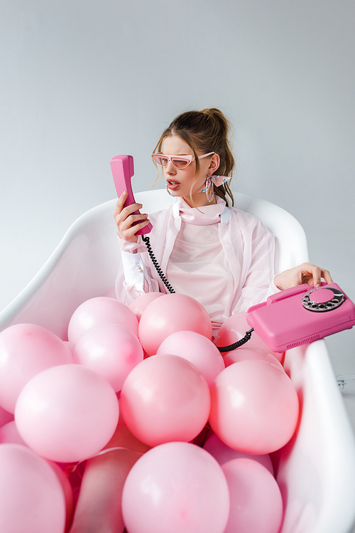 girl in sunglasses looking at retro phone while lying in bathtub with pink air balloons on white