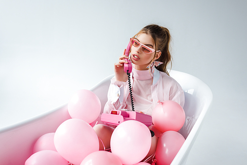 young woman in sunglasses talking on retro phone while lying in bathtub with pink air balloons on white