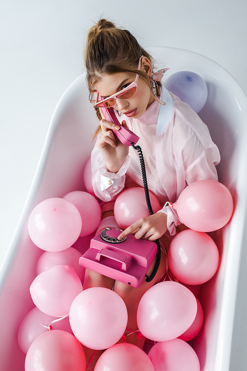 overhead view of girl in sunglasses talking on pink retro phone while lying in bathtub with air balloons on white