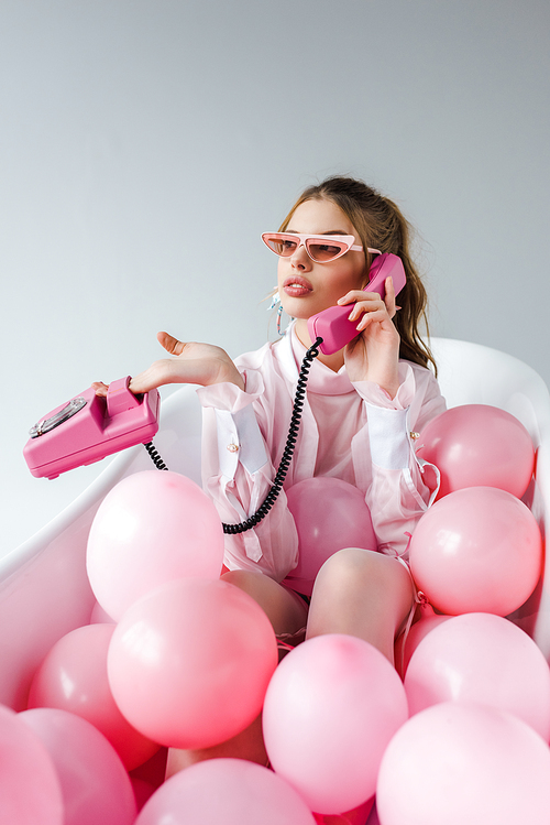 woman in sunglasses talking on retro phone while lying in bathtub with pink air balloons on white