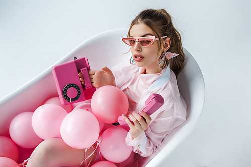 young woman in sunglasses looking at pink retro phone while lying in bathtub with air balloons on white