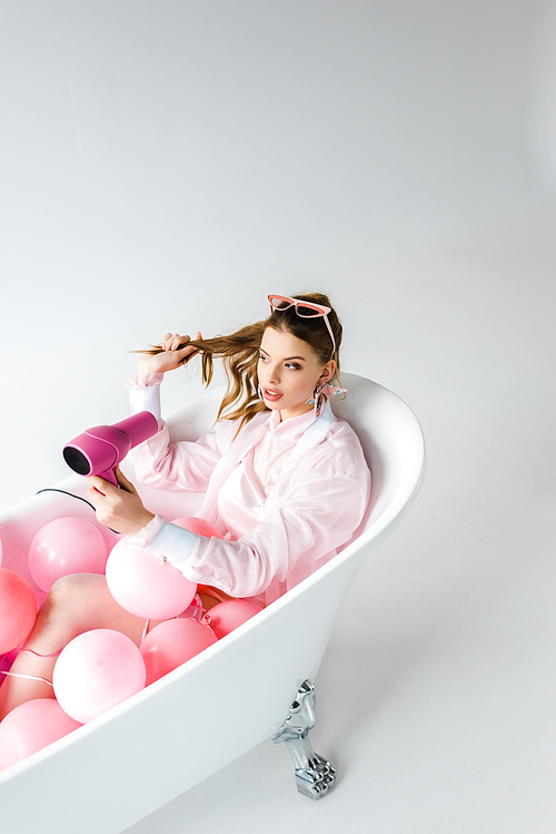 attractive girl using hair dryer while lying in bathtub with pink air balloons on grey