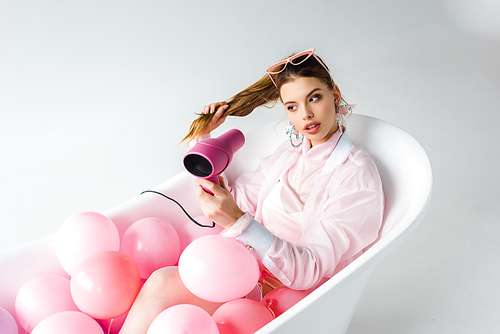attractive girl using hair dryer while lying in bathtub with pink air balloons on white