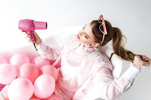 overhead view of girl using hair dryer while lying in bathtub with pink air balloons on white