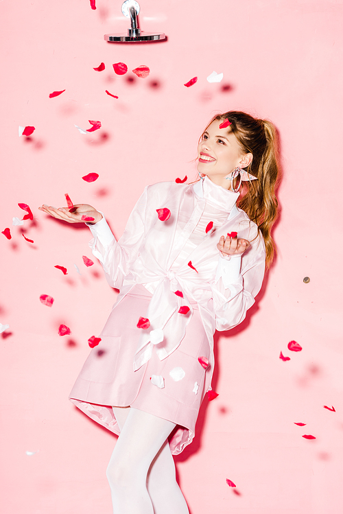 attractive cheerful young woman standing near rose petals on pink