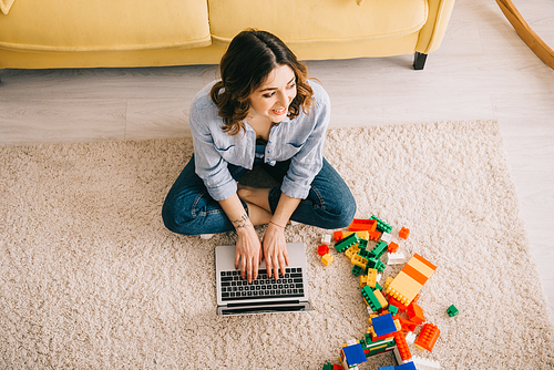 overhead view of smiling woman sitting on carpet near toy blocks and using laptop