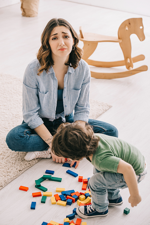 sad tired mother sitting on floor while son playing with toy blocks