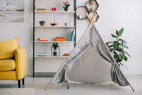 Grey wigwam near shelves with books and yellow sofa