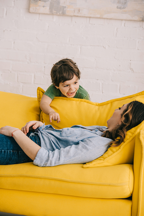 screaming boy standing near tired mother lying on yellow sofa in living room