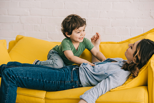 little boy sitting on tired mother while she lying on yellow sofa in living room