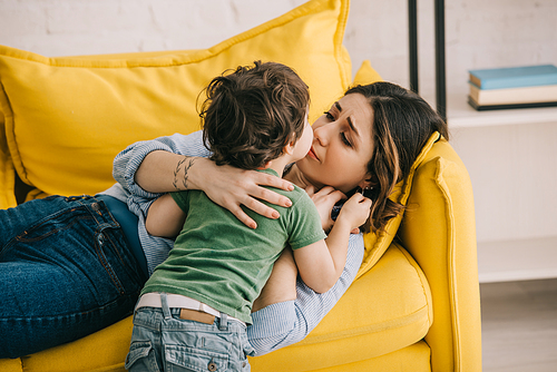 Little boy kissing tired mother lying on yellow sofa